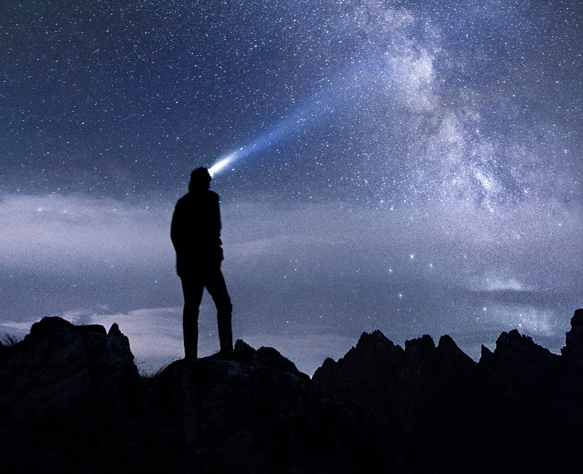 A lone hiker silhouetted on a ridge at night, headlamp beam cutting up into the Milky Way