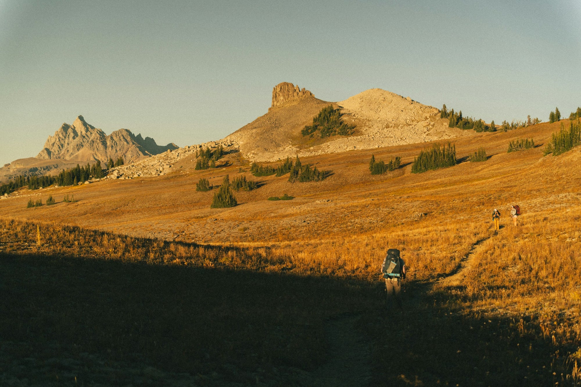 A hiker walking away through a golden meadow below a mountain at sunset