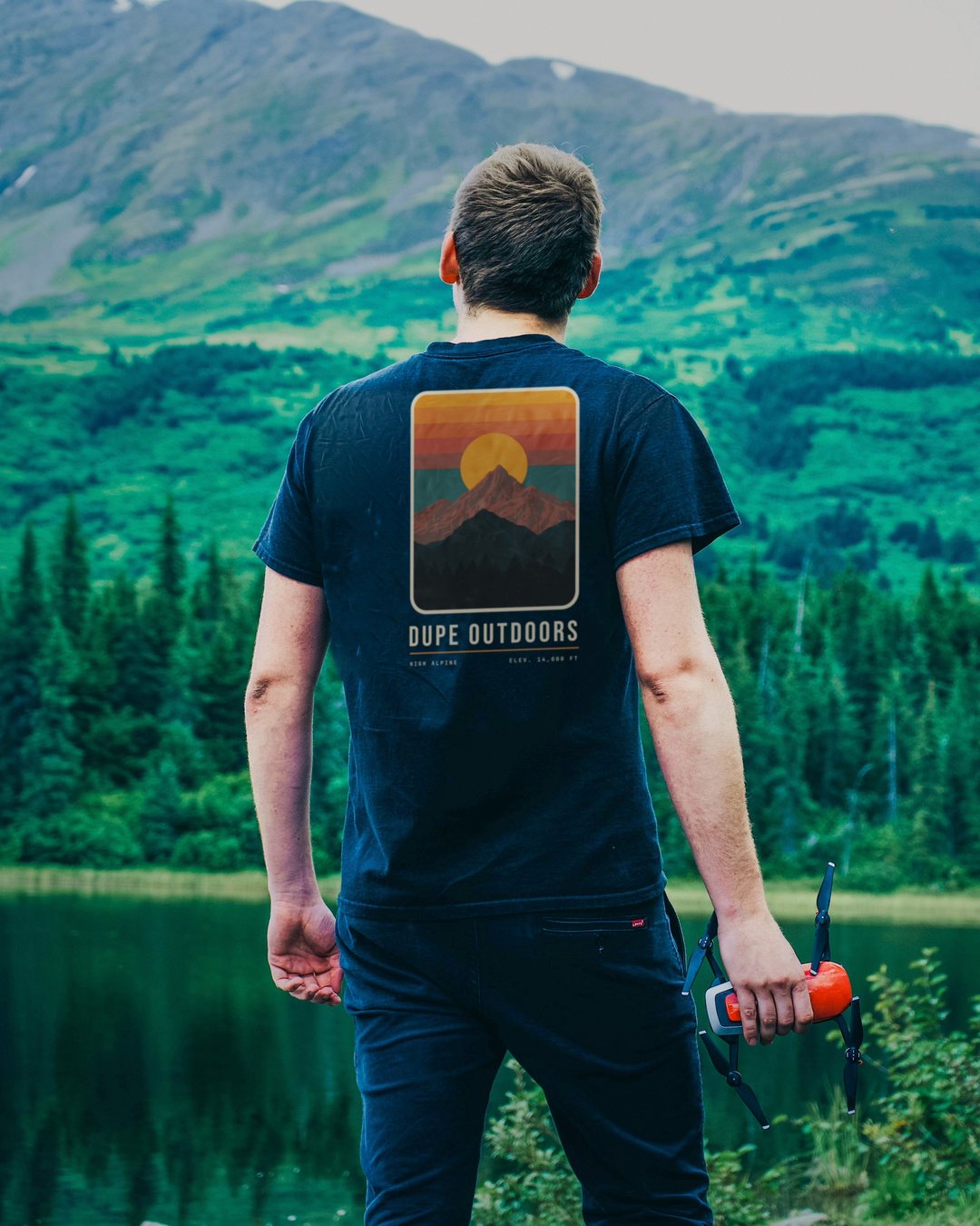 A man at a mountain lake, seen from behind, wearing the Ridgeline tee